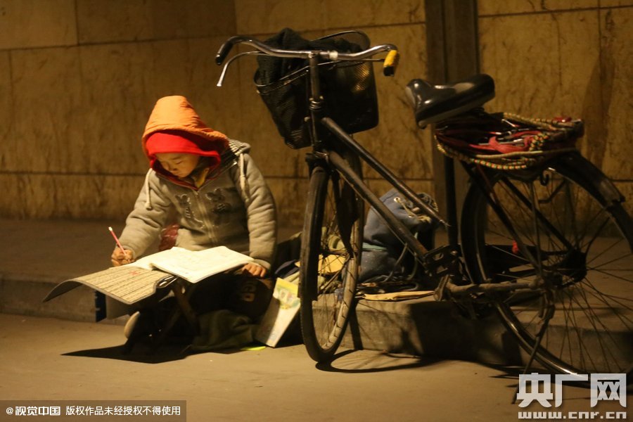 鄭州中原路京廣鐵路大橋下涵洞的馬路牙子邊，一位戴著紅色辣椒帽的小男孩趴在一個(gè)小馬扎上，上面鋪著一張車(chē)墊，在微弱的燈光下寫(xiě)作業(yè)